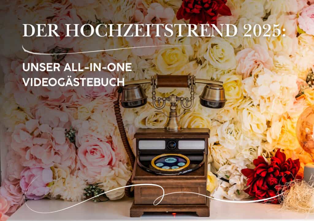 Ein Telefon im Vintage-Stil steht vor einer Wand aus elfenbeinfarbenen, rosa und roten Blumen. Der deutsche Text hebt den Hochzeitstrend 2025 hervor: ein All-in-One-Videogästebuch für unvergessliche Hochzeitserinnerungen.